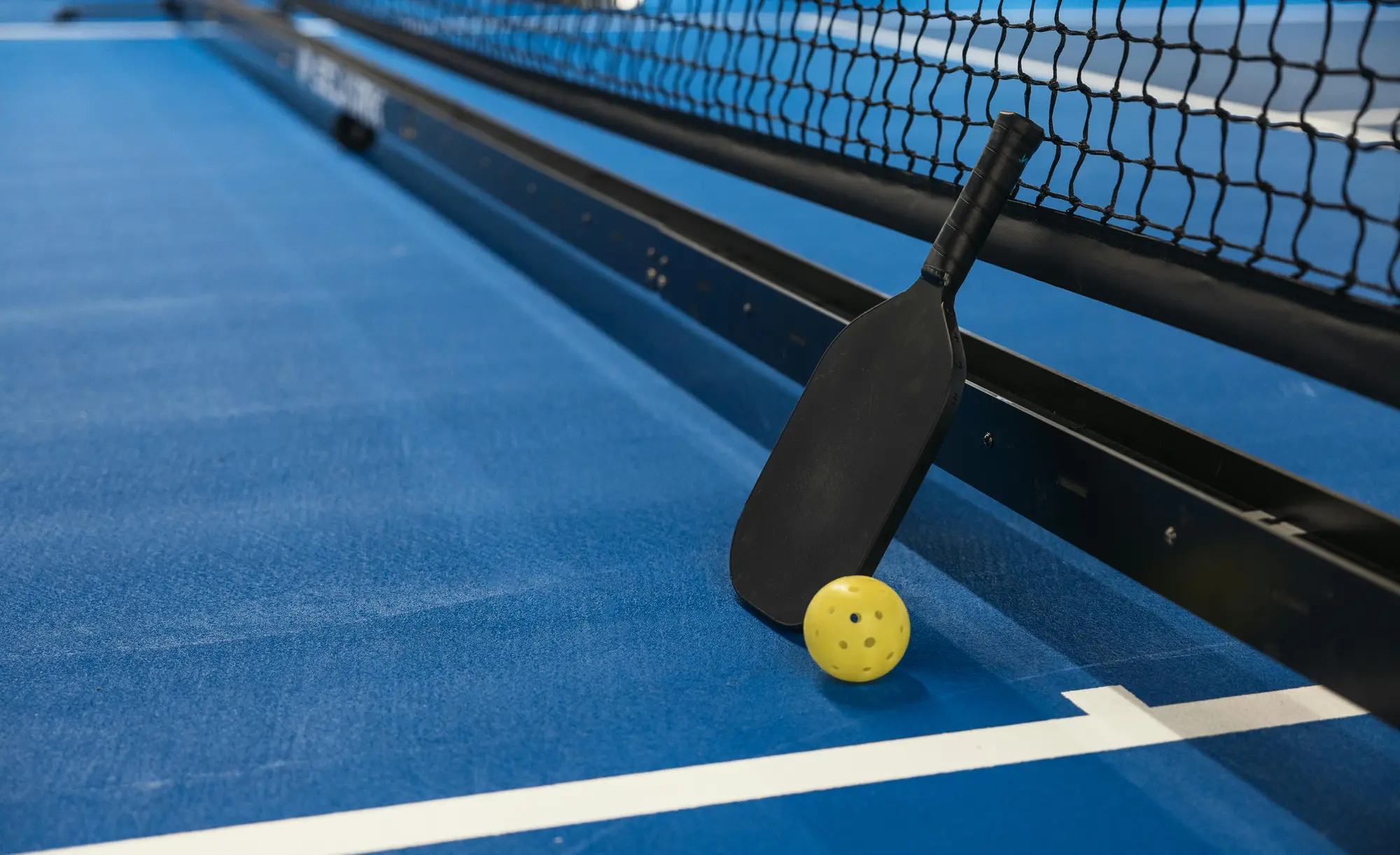 Terrain de pickleball intérieur bleu avec raquette noire et balle jaune sur la ligne, vu en plongée