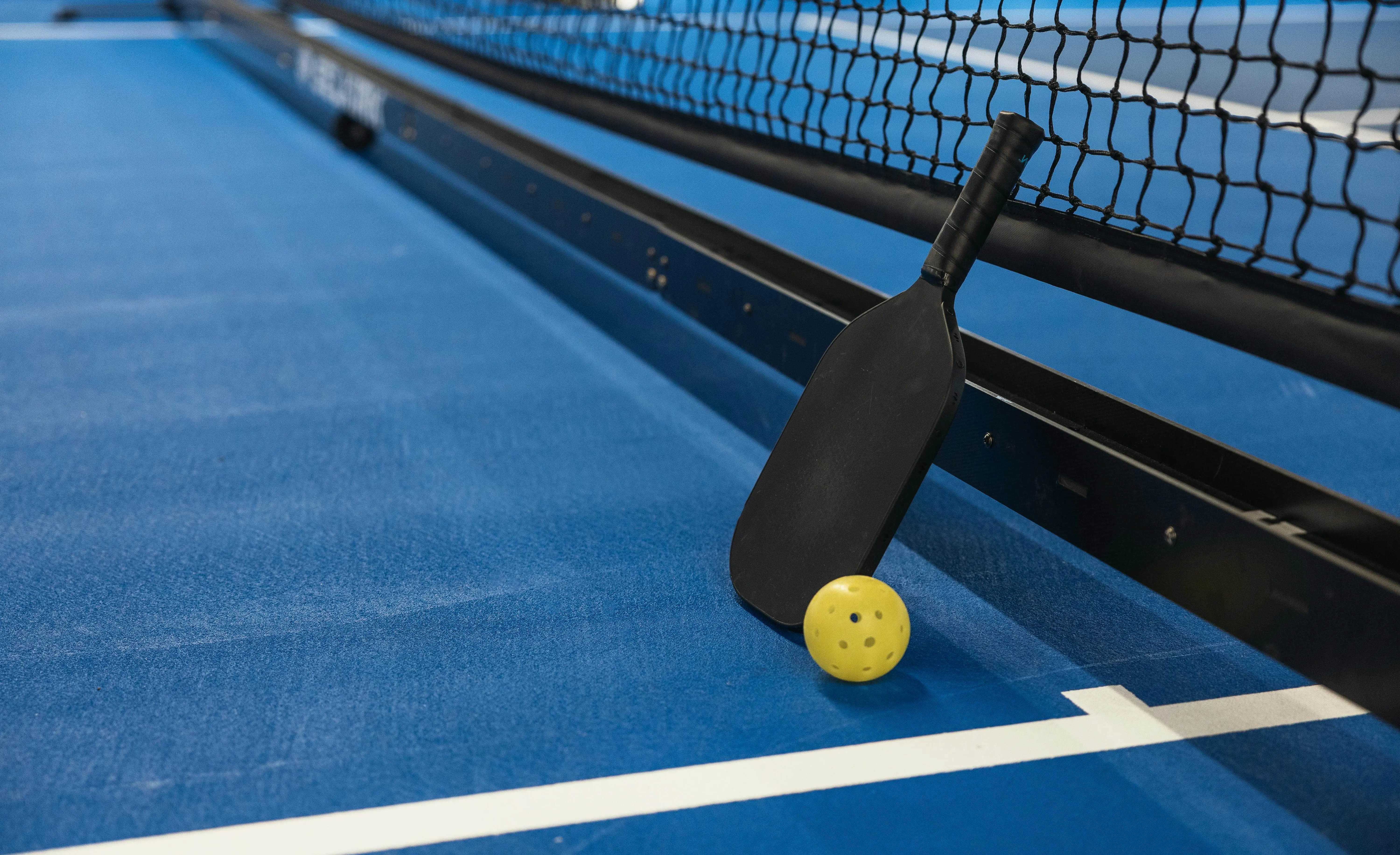 Terrain de pickleball intérieur bleu avec raquette noire et balle jaune sur la ligne, vu en plongée