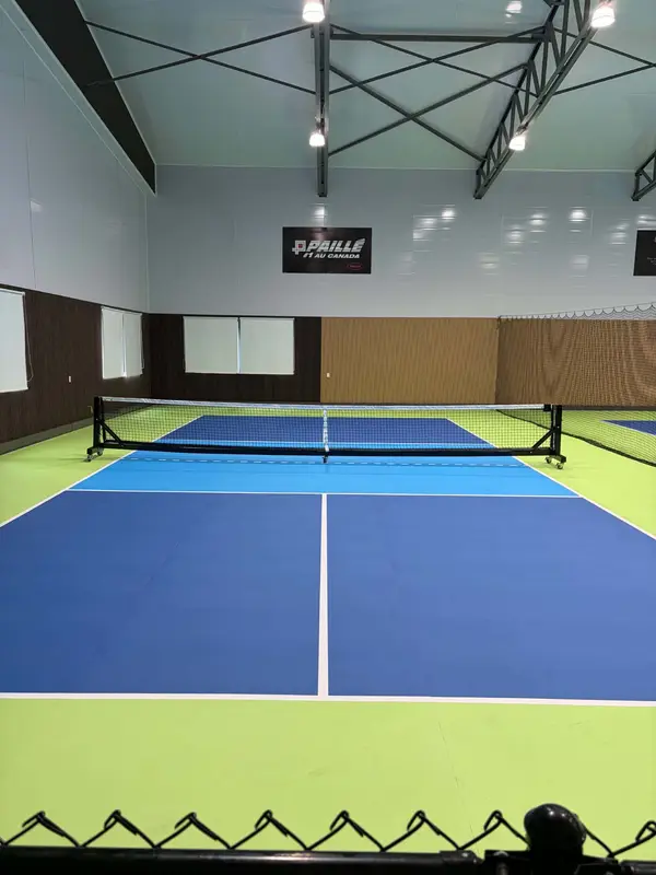 Terrain de pickleball bleu et vert, enseigne Paillé au mur du fond, fenêtres sur le parcours de golf à gauche