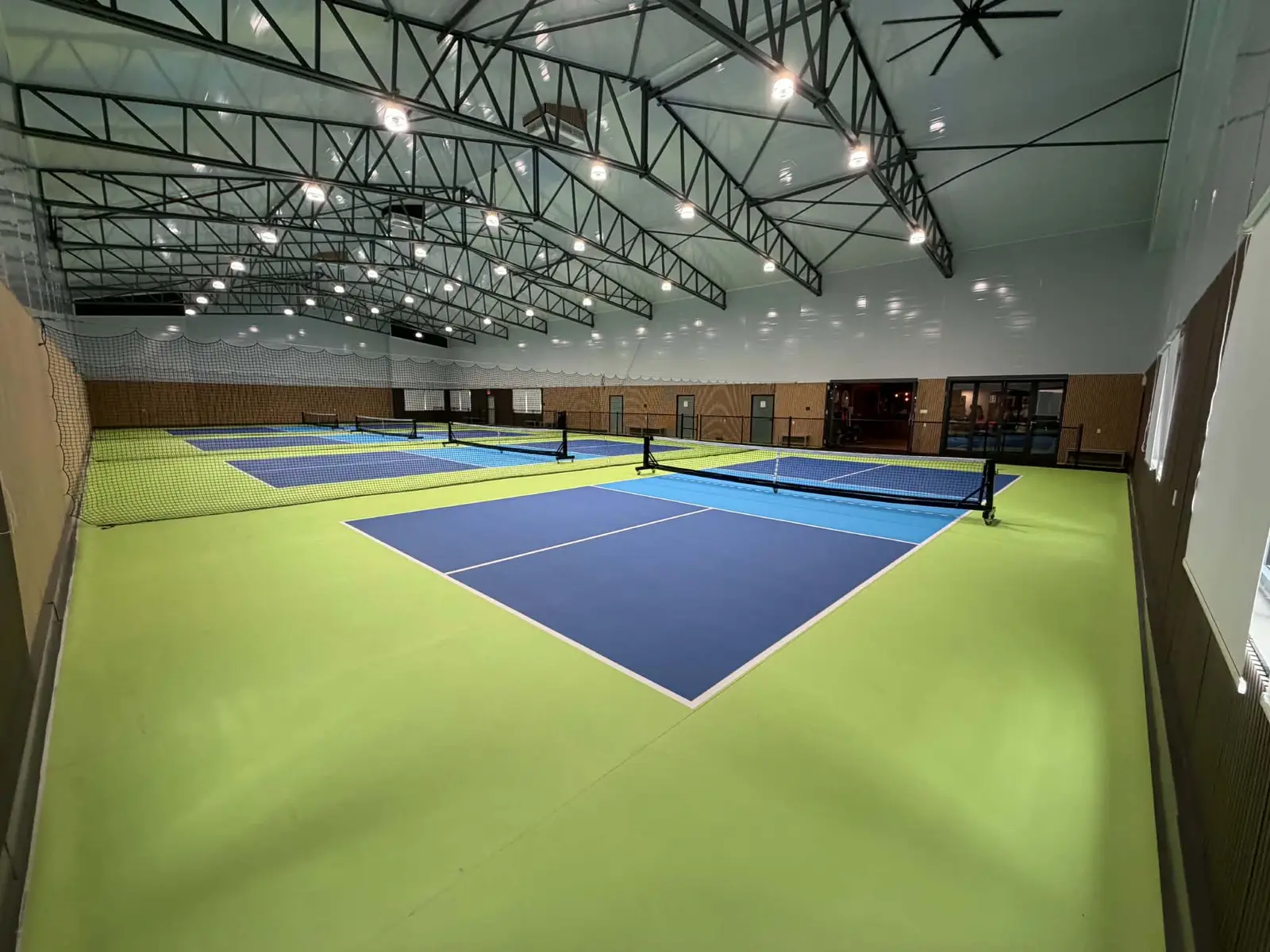 Vue d'ensemble du pavillon : les quatre terrains de pickleball bleu et vert sous la charpente d'acier, avec les portes vitrées du club house au fond